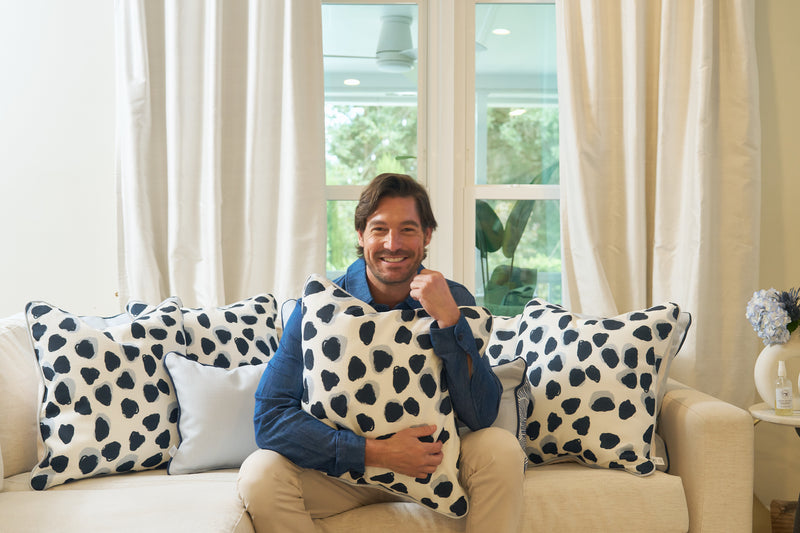 A man sits on a beige sofa, smiling and holding an Oversized Modern Polka Dots Pillow with a hand-painted pattern. Similar black-spotted pillows line the sofa, enhancing the southern decor. White curtains frame a window overlooking greenery.
