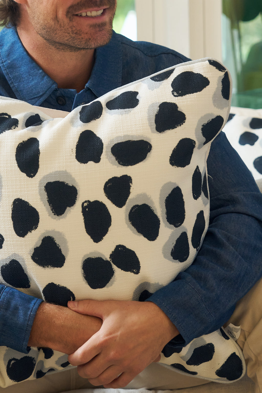 A person in a blue shirt sits holding an Oversized Modern Polka Dots Pillow, which features black and gray irregular spots for a hand-painted look inspired by southern decor.