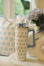 The Mini Calico 30oz Tumbler, featuring a handle, straw, and blue sunburst pattern, sits on a white surface beside a vase of blue hydrangeas and a cushion with a matching sunburst design.