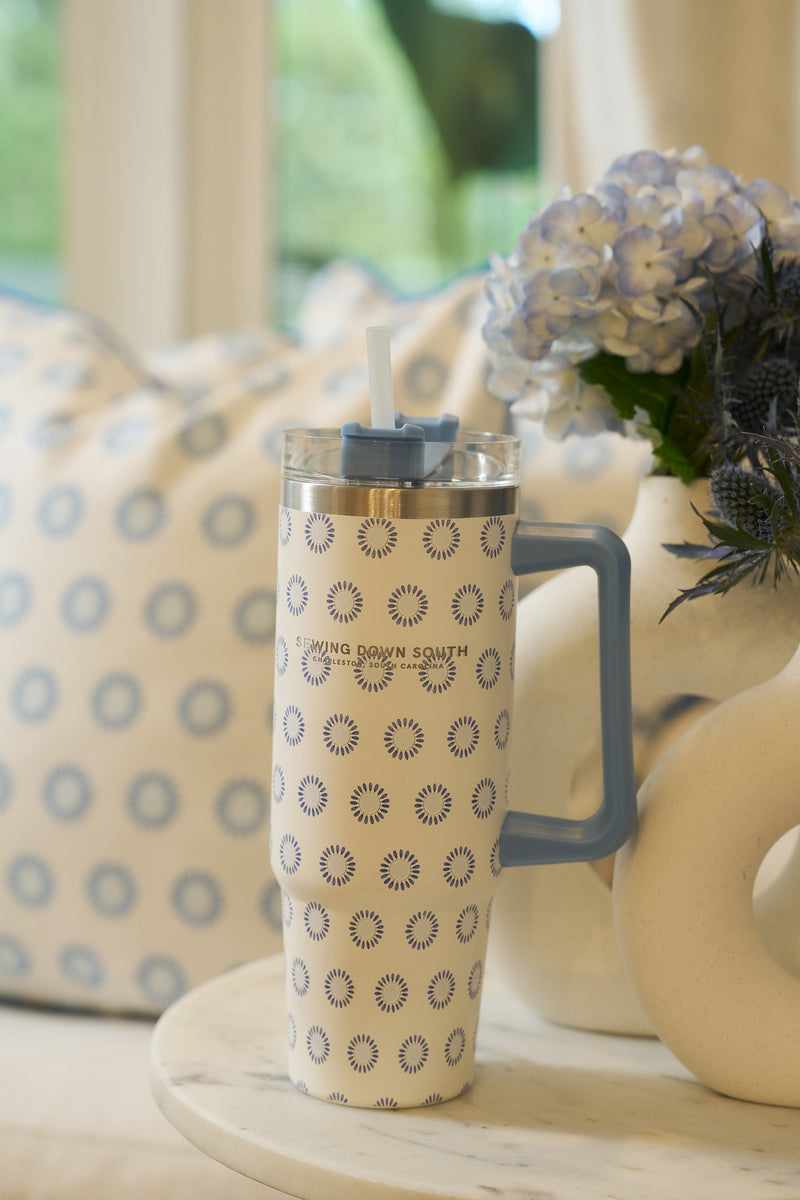 The Mini Calico 30oz Tumbler, featuring a handle, straw, and blue sunburst pattern, sits on a white surface beside a vase of blue hydrangeas and a cushion with a matching sunburst design.
