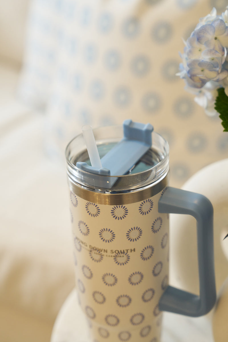A Mini Calico 30oz Tumbler with a blue handle and lid, plastic straw, and blue circular pattern rests on a white surface. Its no-sweat finish shines, with a pillow and blue hydrangea flower softly blurred in the background.