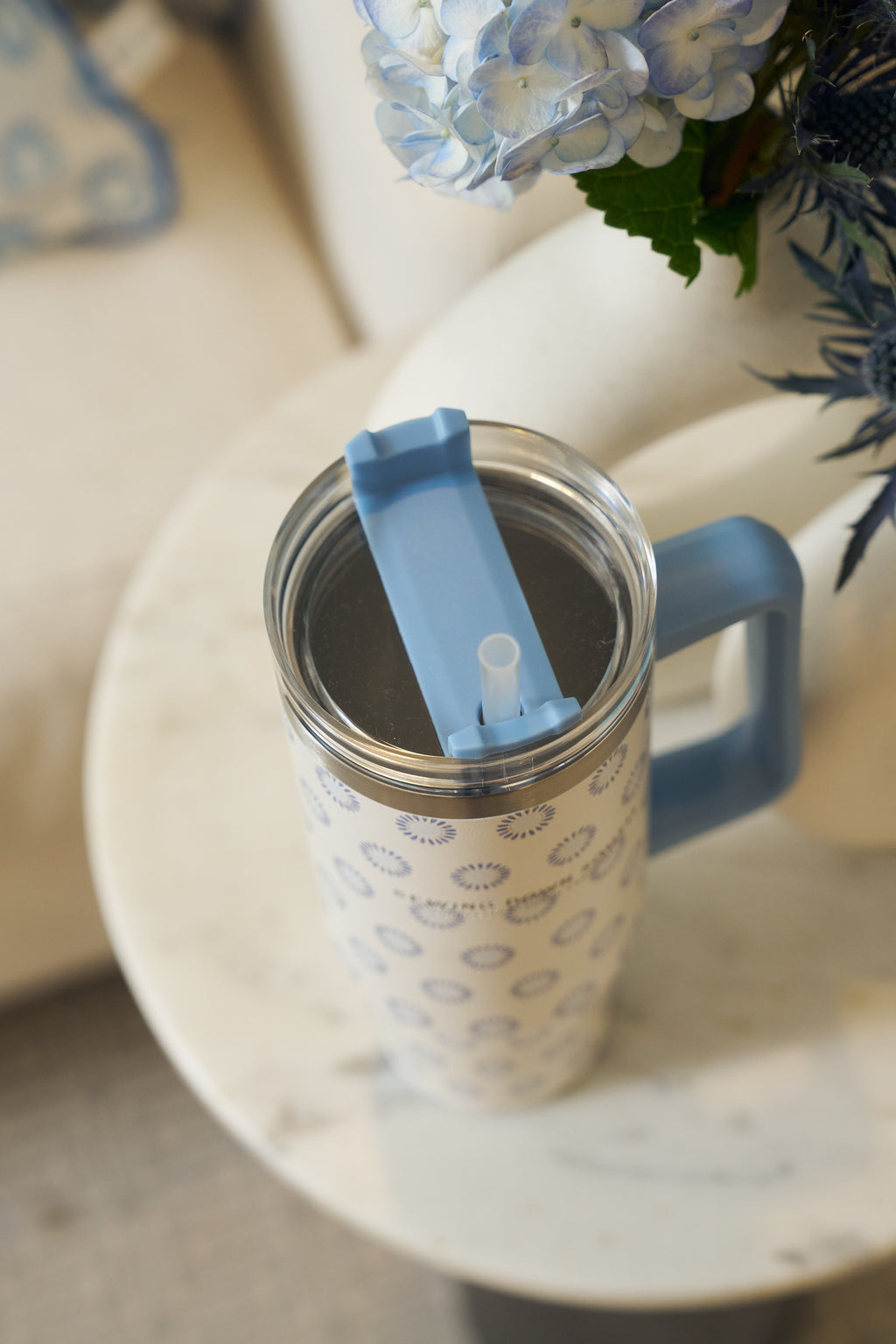 The Mini Calico 30oz Tumbler, featuring a patterned stainless steel design with a blue handle and flip-top lid, rests on a round white marble table beside a vase of blue and white hydrangeas.