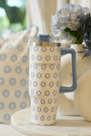 The Mini Calico 30oz Tumbler, featuring a blue handle, blue and white circular pattern, and straw, sits on a round white table. Its insulated design keeps drinks cool beside a pillow and a vase of blue flowers in the background.