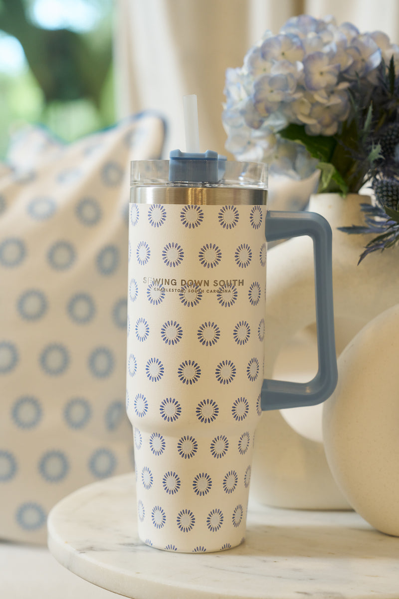 The Mini Calico 30oz Tumbler, featuring a blue handle, blue and white circular pattern, and straw, sits on a round white table. Its insulated design keeps drinks cool beside a pillow and a vase of blue flowers in the background.