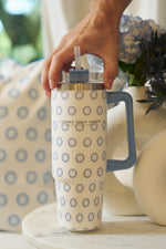 A hand holds the Mini Calico 30oz Tumbler with a blue handle and repeating blue circular pattern, beside a vase of blue and white flowers on a marble table. Part of the Sewing Down South collection.