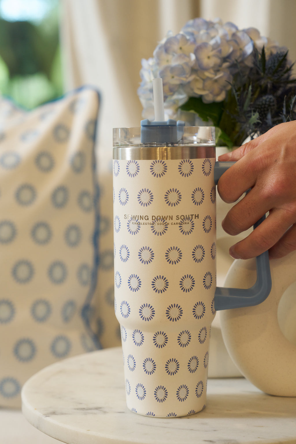 A hand holds the Mini Calico 30oz Tumbler—tall, patterned stainless steel with a blue handle and straw—on a marble table. In the background are a pillow with matching circular design and a vase of blue hydrangeas.