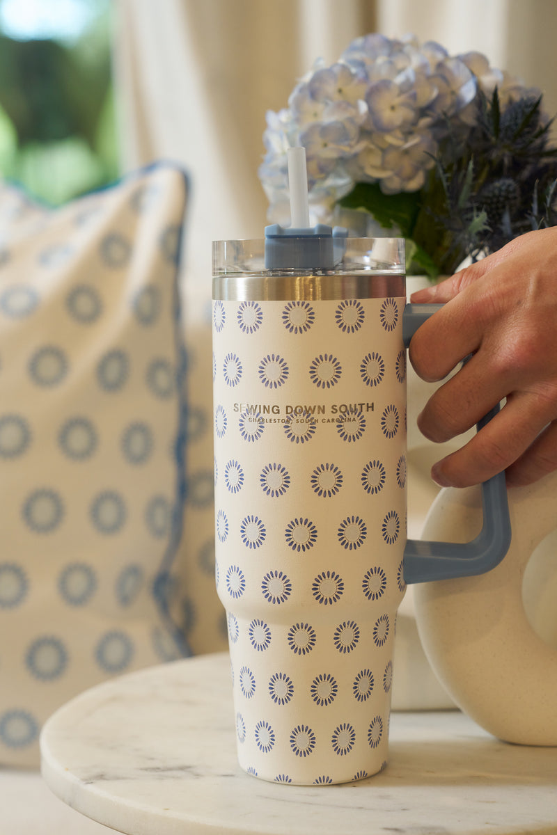 A hand holds the Mini Calico 30oz Tumbler—tall, patterned stainless steel with a blue handle and straw—on a marble table. In the background are a pillow with matching circular design and a vase of blue hydrangeas.