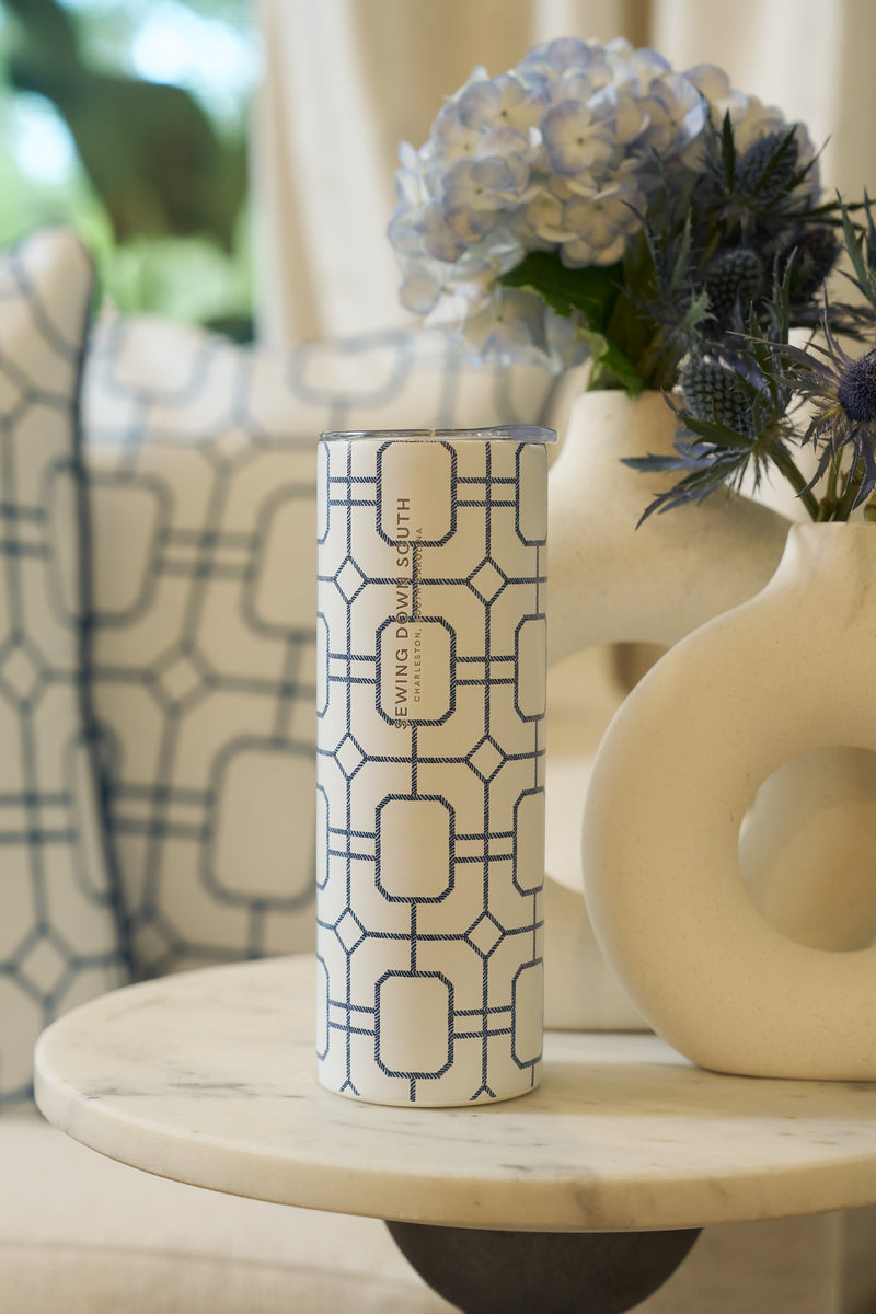 A Blue Lattice 20oz Tumbler from Sewing Down South rests on a marble table beside a cream vase with blue and white flowers, with a cushion featuring a matching geometric design in the background.