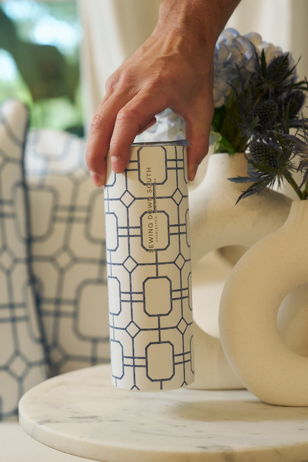 A hand holds the Blue Lattice 20oz Tumbler from Sewing Down South, a stainless steel insulated tumbler with a blue geometric pattern and EVINO & SMITH label, resting on marble next to a white vase with flowers.