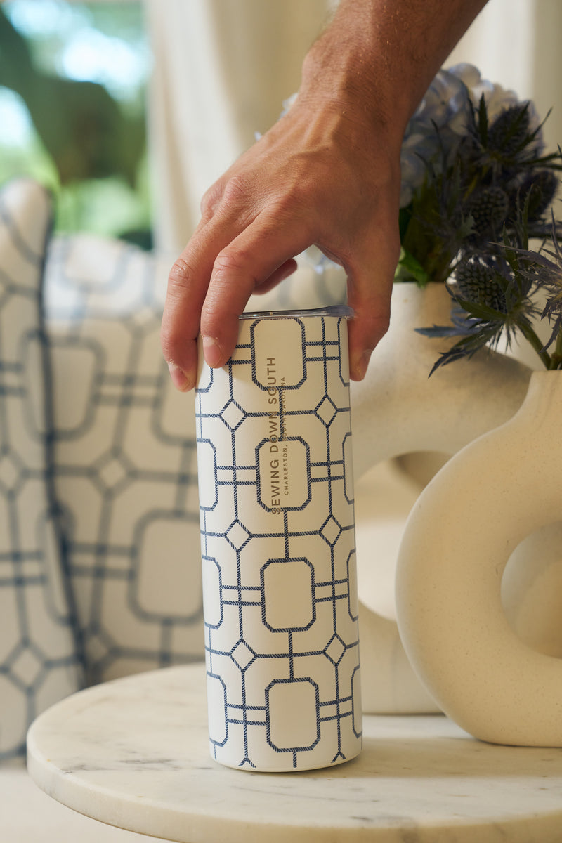 A hand holds a Blue Lattice 20oz Tumbler on a round marble table, with abstract decor and flowers softly blurred in the background—a stylish nod to Sewing Down South.