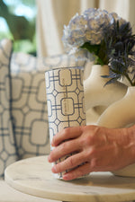 A hand holds the Blue Lattice 20oz Tumbler on a marble table, with a vase of blue and purple flowers and a geometric-patterned pillow in the background.