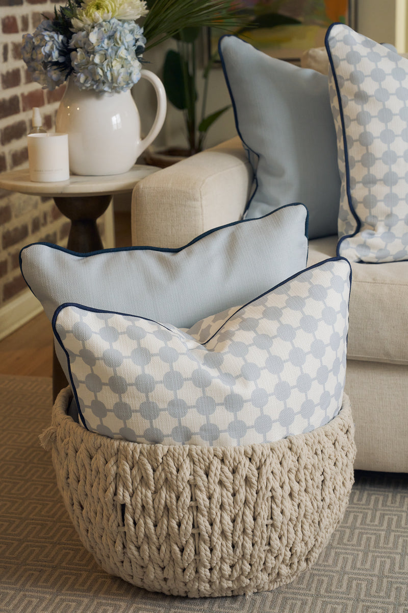 An Oversized Mod Dot Grid Pillow in light blue and white with hand-drawn patterns sits in a chunky knitted beige basket by a cream sofa, beside a side table with a pitcher and flowers—a modern Southern decor accent.