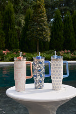 A Pink Lattice 30oz Tumbler with handle and straw sits on a white table by the pool, featuring a no sweat exterior. Tall green trees and pink flowers provide a vibrant background.