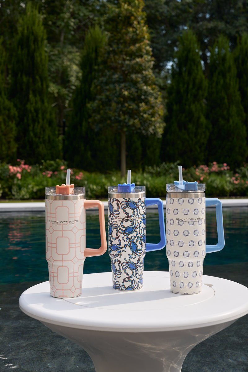 A Pink Lattice 30oz Tumbler with handle and straw sits on a white table by the pool, featuring a no sweat exterior. Tall green trees and pink flowers provide a vibrant background.