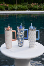 Three Crab Craze 30oz Tumblers with handles and straws rest on a white round table by the pool, their insulated stainless steel ensuring a no-sweat exterior, surrounded by greenery and blooming flowers.