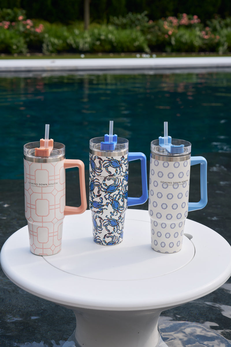 Three Crab Craze 30oz Tumblers with handles and straws rest on a white round table by the pool, their insulated stainless steel ensuring a no-sweat exterior, surrounded by greenery and blooming flowers.