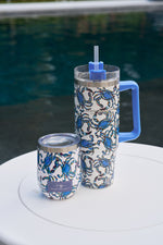 A Crab Craze 30oz Tumbler with handle and straw, plus a shorter matching cup—both with blue crab designs and no-sweat exteriors—rest on a round white table by the pool.