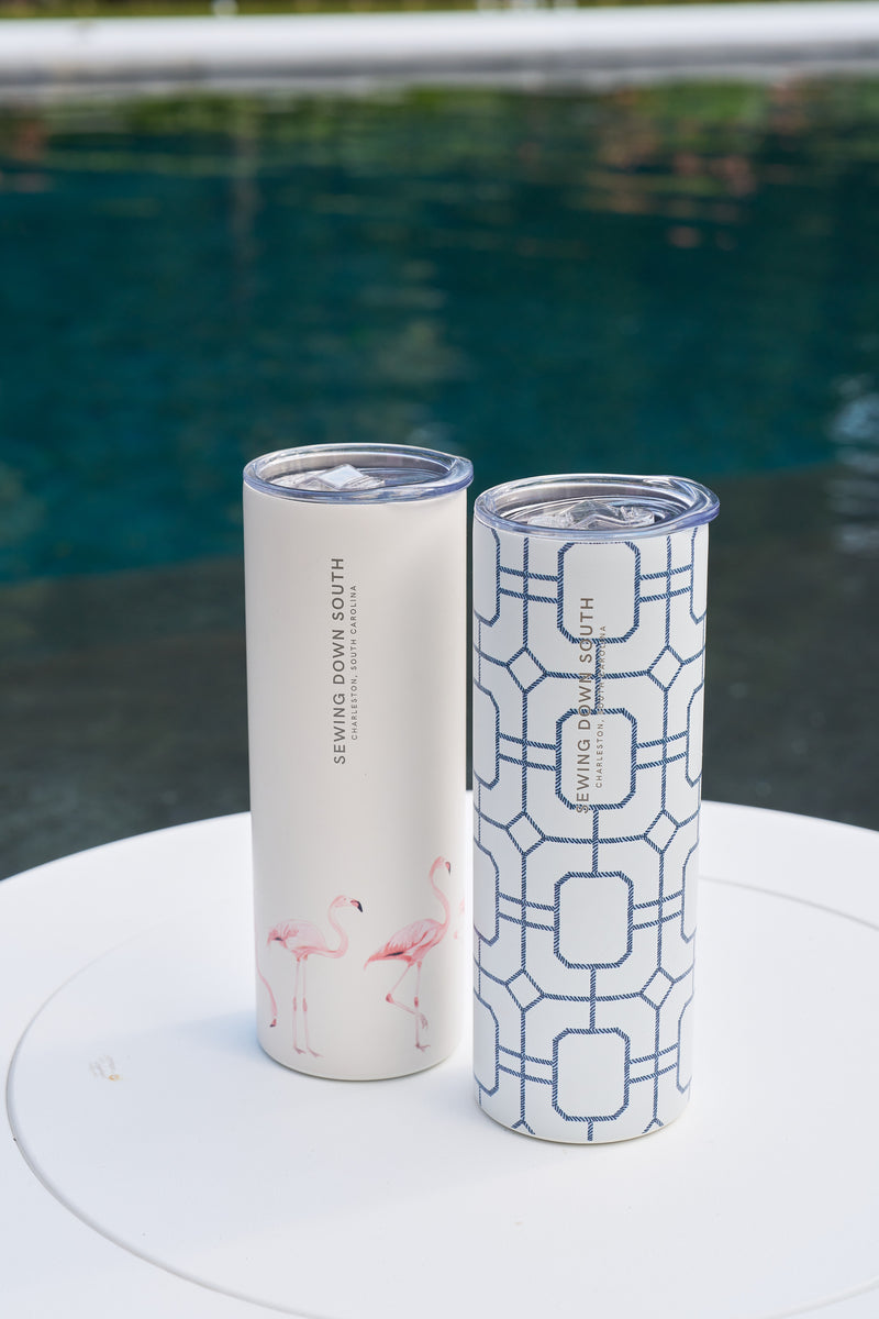 Two tall stainless steel tumblers with lids sit poolside. One 20oz cup is cream with pink flamingos, the other is the Blue Lattice 20oz Tumbler, white with a blue geometric pattern. Both have a no sweat exterior and SEWING DOWN SOUTH label.