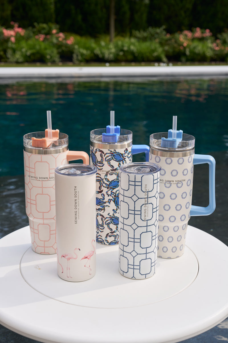 Five Blue Lattice 20oz stainless steel tumblers with straws and no-sweat exterior are arranged on a white round table beside a swimming pool, with greenery and flowers in the background.