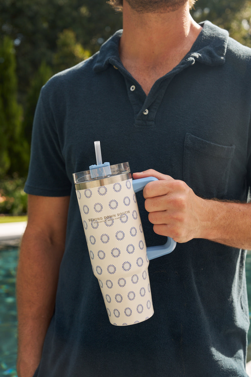 A person in a dark blue shirt holds the Mini Calico 30oz Tumbler, a large patterned stainless steel cup with handle and straw. The no sweat exterior stands out against blurred greenery and a swimming pool in the background.