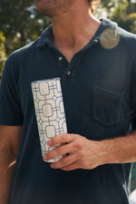A person in a dark blue collared shirt holds the Blue Lattice 20oz Tumbler with a geometric blue pattern and beige label. Its no sweat exterior keeps hands dry as sunlight and greenery brighten the background.