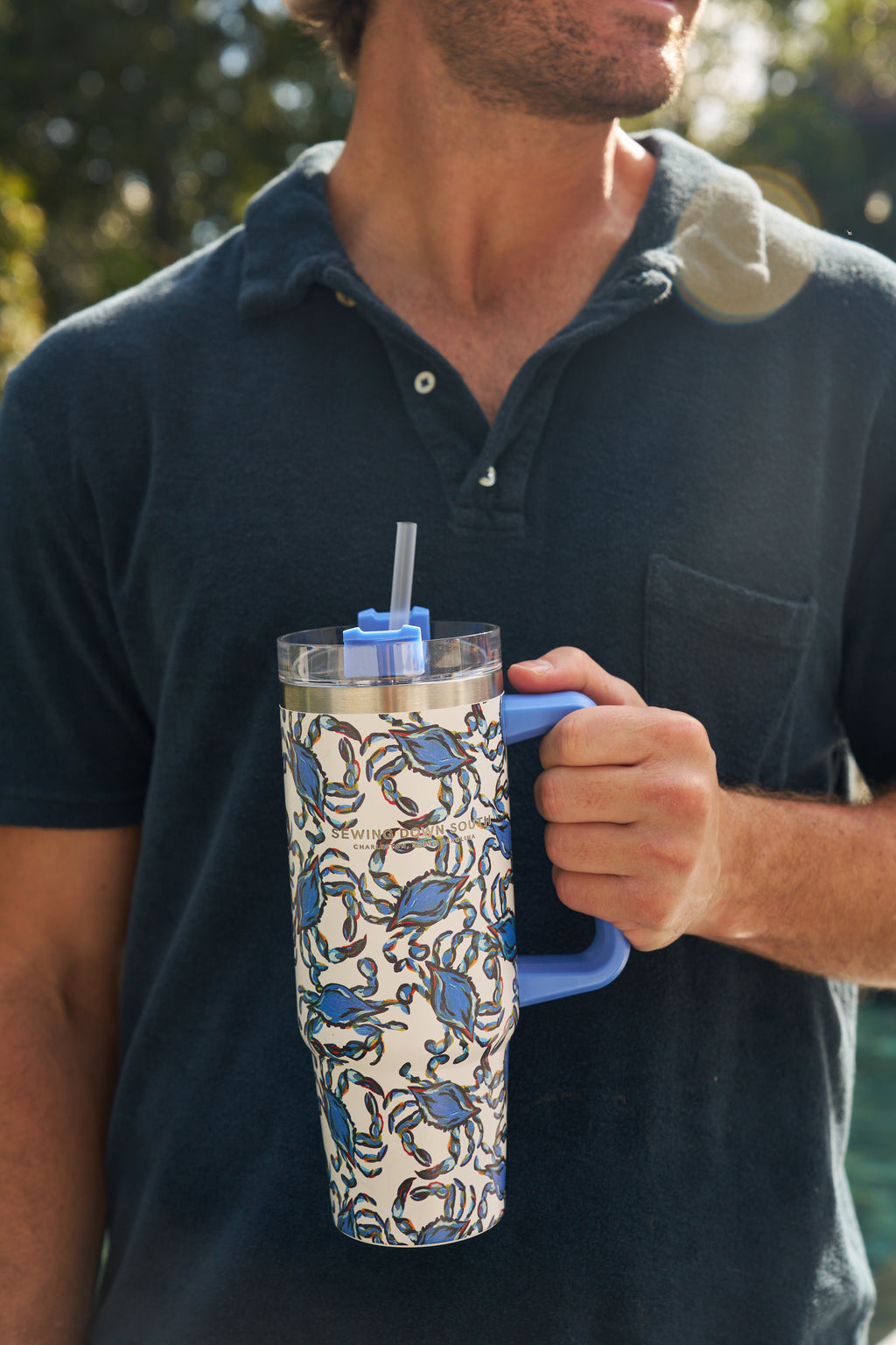 A man in a dark blue polo shirt holds the Crab Craze 30oz Tumbler, a stainless steel insulated cup with a blue handle, blue crab design, plastic straw, and no-sweat exterior—ideal for enjoying drinks outdoors.