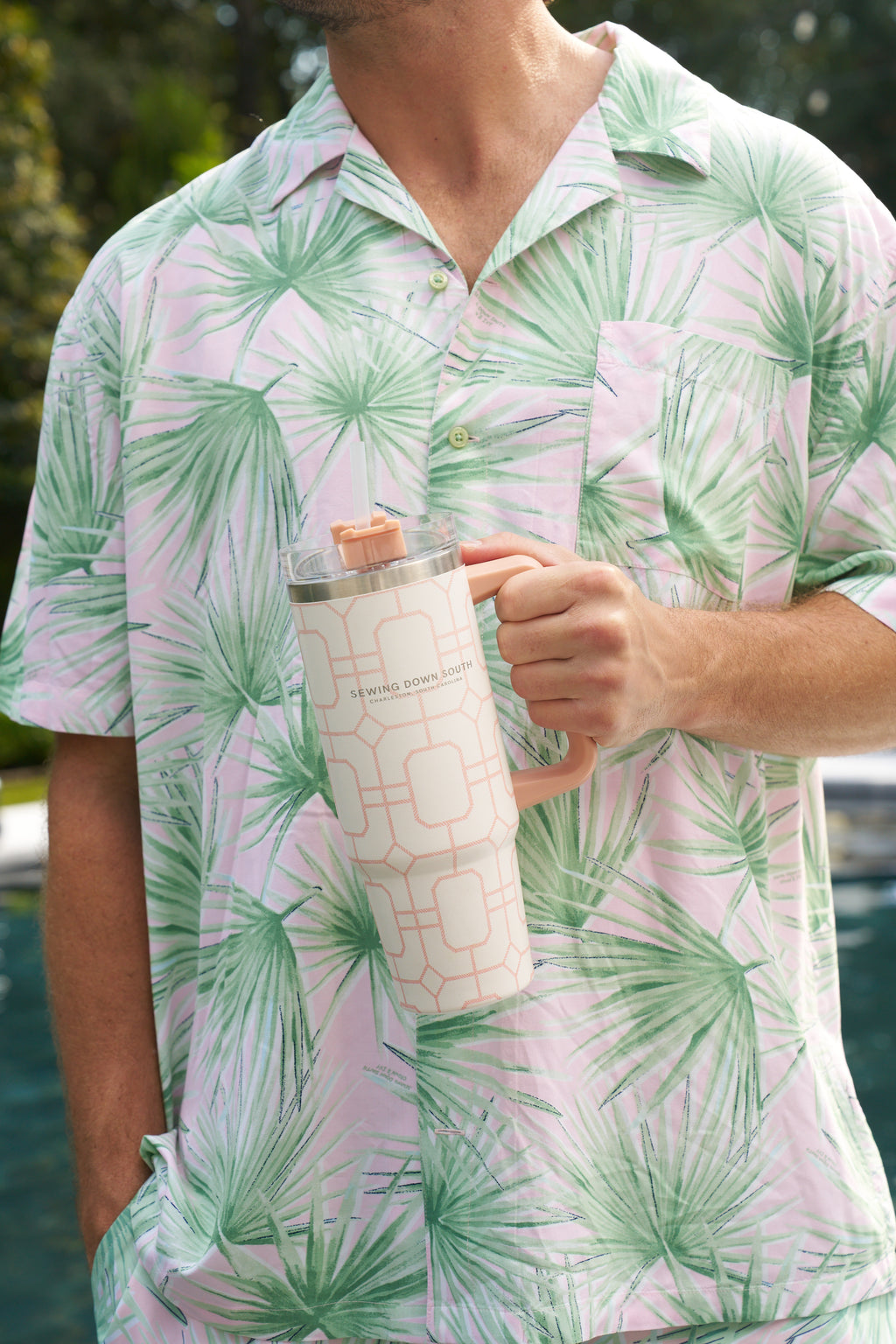 A person in a light pink and green palm leaf shirt stands by a pool, holding a Pink Lattice 30oz Tumbler with a no sweat exterior, lid, and straw. Their other hand is casually resting in their pocket.