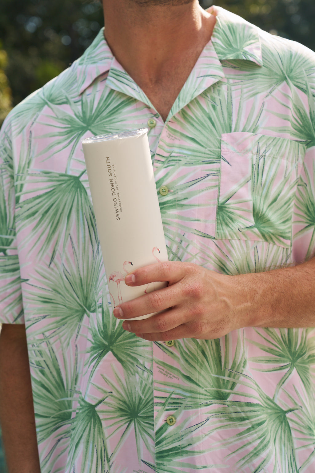 Someone wearing a pink and green palm leaf shirt holds the Flamingo 20oz Tumbler with a no sweat exterior, standing outdoors in natural light.