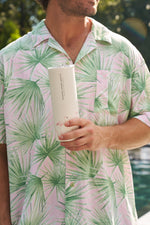 A man outdoors, wearing a pink and green palm-leaf shirt, holds the Flamingo 20oz Tumbler with flamingo prints near his chest; the blurred background suggests a pool and greenery.