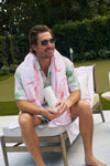 A man with short brown hair and sunglasses relaxes outdoors in a tropical shirt, holding a Flamingo 20oz Tumbler with a no sweat exterior, sitting on a lounge chair near greenery and a putting green.