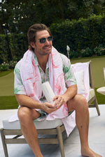 A man with short brown hair and sunglasses relaxes outdoors in a tropical shirt, holding a Flamingo 20oz Tumbler with a no sweat exterior, sitting on a lounge chair near greenery and a putting green.
