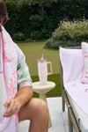 A person with a pink and green towel holds the Pink Lattice 30oz Tumbler by an outdoor sofa, while another tumbler with a straw sits on a beige table nearby—both featuring a no sweat exterior in the garden setting.
