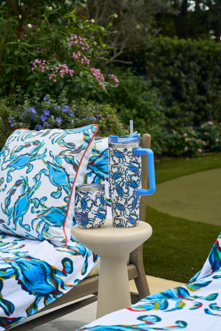A patio scene with a cushioned lounge chair and small table holding the Crab Craze Shades of Blue Wine Tumbler. Blue swirl cushions and towels, plus blooming flowers and green plants, complete the coastal-inspired setting.