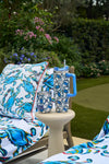 A patio scene with a cushioned lounge chair and small table holding the Crab Craze Shades of Blue Wine Tumbler. Blue swirl cushions and towels, plus blooming flowers and green plants, complete the coastal-inspired setting.