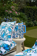 A patio scene with a cushioned lounge chair and small table holding the Crab Craze Shades of Blue Wine Tumbler. Blue swirl cushions and towels, plus blooming flowers and green plants, complete the coastal-inspired setting.