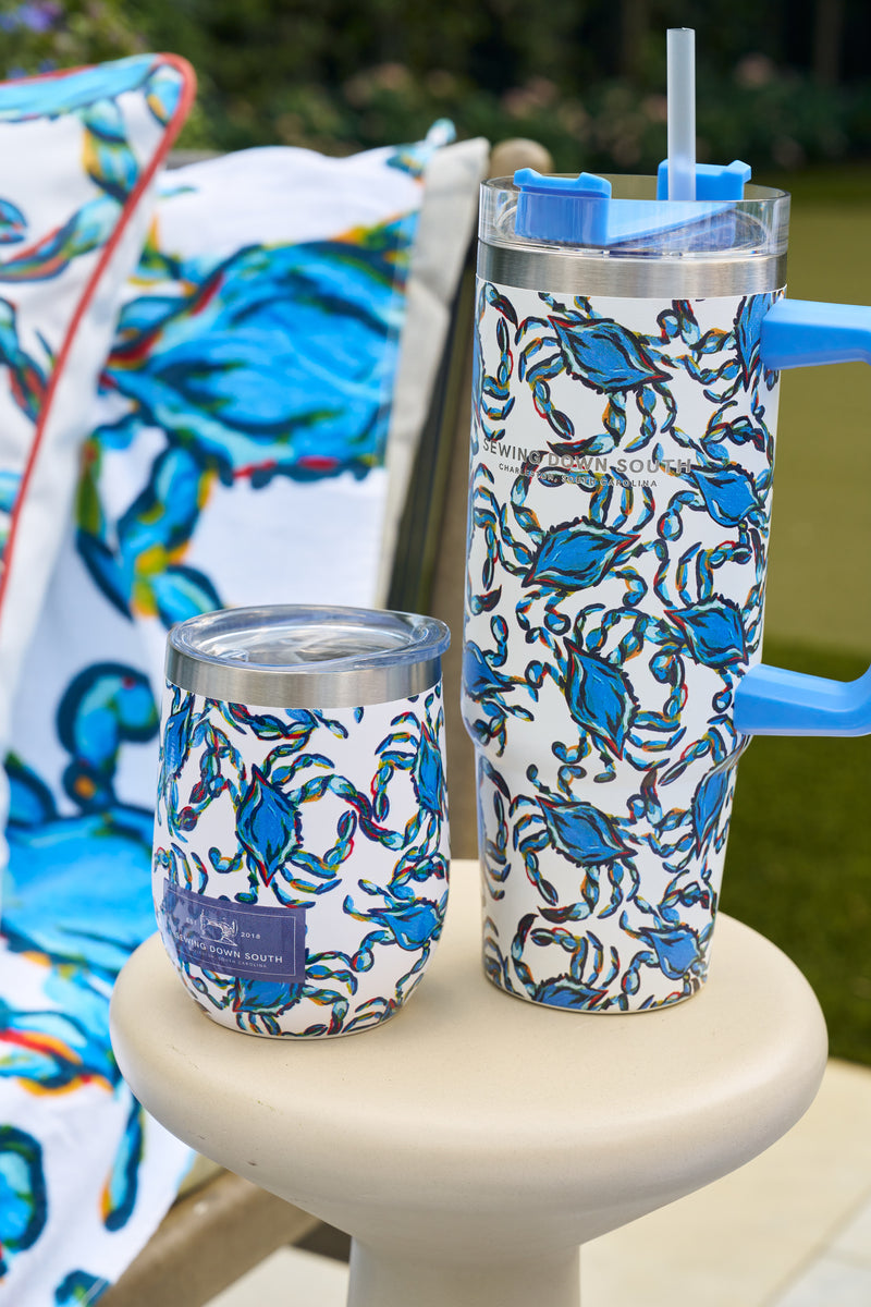 Two Crab Craze 30oz Tumblers with blue crab designs and no sweat exteriors rest on a round outdoor table, while a matching crab-patterned towel hangs over a nearby chair.