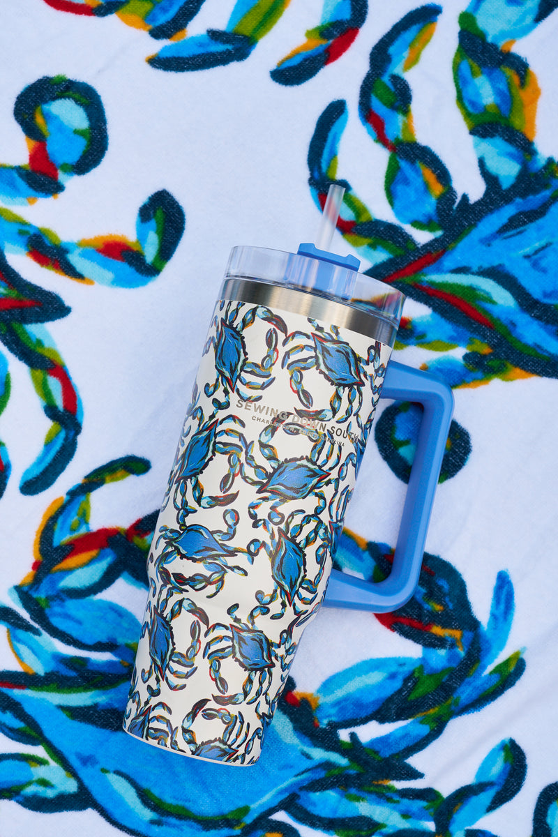 The Crab Craze 30oz Tumbler with a blue handle, straw, and blue crab pattern from Sewing Down South sits on a matching vibrant towel.