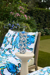 The Crab Craze 30oz Tumbler with a blue straw sits on a white table next to a chair draped with a Sewing Down South towel, surrounded by green plants and purple flowers in the garden.