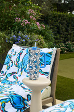 The Crab Craze 30oz Tumbler with a blue straw sits on a white table next to a chair draped with a Sewing Down South towel, surrounded by green plants and purple flowers in the garden.