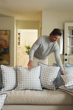 A smiling man in a light gray sweater arranges the Oversized Mod Dot Grid Pillow, featuring a hand-drawn design, on a light sofa in a bright, modern Southern-style living room.