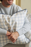 A person wearing a light gray sweater sits on a sofa, smiling and holding the Oversized Mod Dot Grid Pillow, which features a hand-drawn pattern. Only their hands and lower face are shown, showcasing modern Southern decor style.