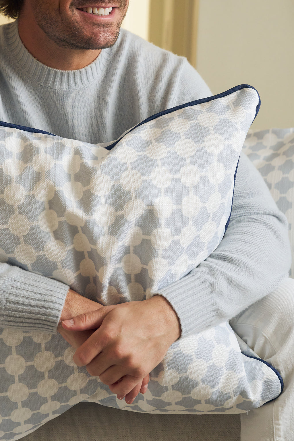 A person wearing a light gray sweater sits on a sofa, smiling and holding the Oversized Mod Dot Grid Pillow, which features a hand-drawn pattern. Only their hands and lower face are shown, showcasing modern Southern decor style.
