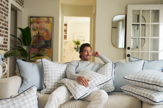 A man with dark hair and a beard sits on a couch surrounded by light blue and white patterned pillows, smiling and resting his arm on the back of the sofa in a cozy, well-decorated living room.