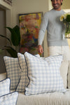 A man in a light blue sweater and white pants holds a vase of flowers behind a beige sofa adorned with the Oversized Mod Dot Grid Pillow, adding modern Southern decor to the stylish living room.
