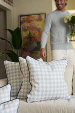A man in a light blue sweater and white pants holds a vase of flowers behind a beige sofa adorned with the Oversized Mod Dot Grid Pillow, adding modern Southern decor to the stylish living room.