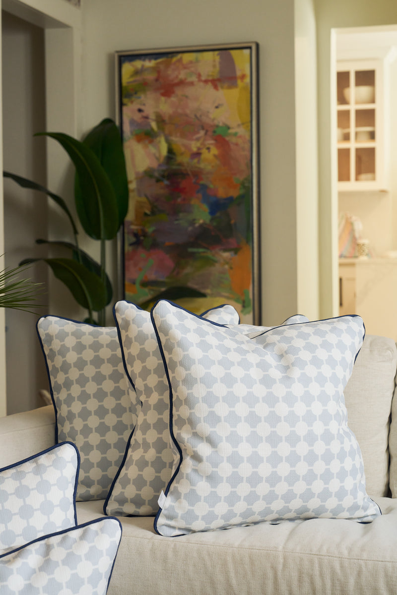 An Oversized Mod Dot Grid Pillow with a hand-drawn pattern is arranged among three decorative pillows on a beige sofa, complemented by a large abstract painting and a leafy plant for modern Southern style.