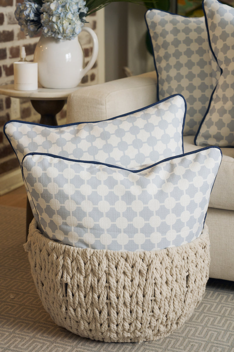 A woven basket holds two Oversized Mod Dot Grid Pillows with a hand-drawn exclusive pattern beside a beige sofa with matching pillows. A side table topped with candles and flowers completes the modern Southern look.