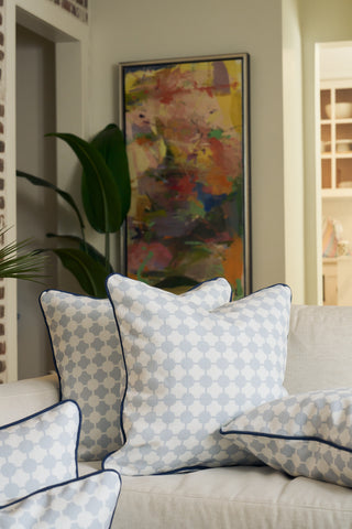 A beige sofa with blue and white Oversized Mod Dot Grid Pillows—featuring an exclusive hand-drawn pattern—sits in front of a houseplant and colorful abstract art, creating a cozy, modern Southern look.