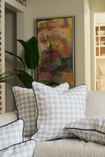 A beige sofa with blue and white Oversized Mod Dot Grid Pillows—featuring an exclusive hand-drawn pattern—sits in front of a houseplant and colorful abstract art, creating a cozy, modern Southern look.