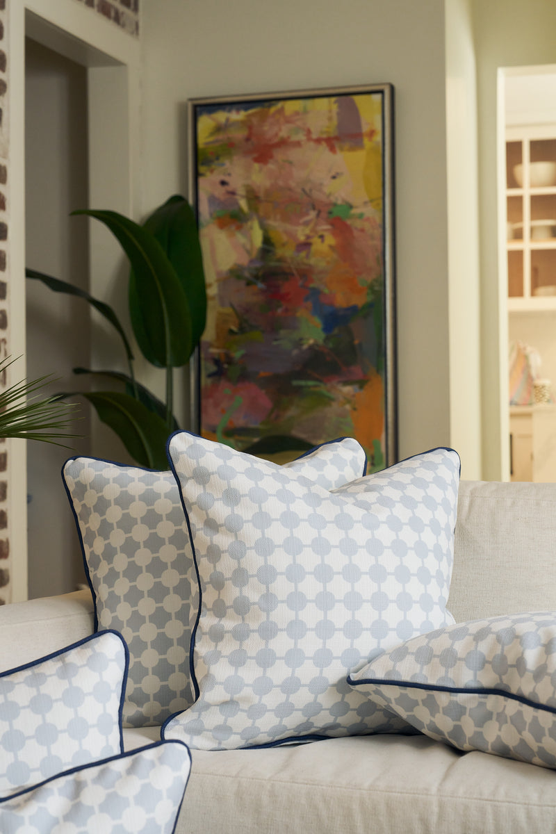 A beige sofa with blue and white Oversized Mod Dot Grid Pillows—featuring an exclusive hand-drawn pattern—sits in front of a houseplant and colorful abstract art, creating a cozy, modern Southern look.